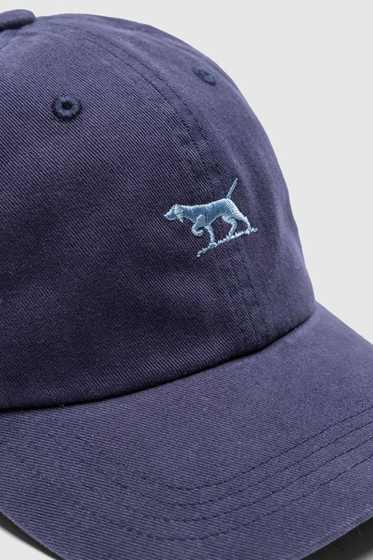 A close-up of the Rodd & Gunn Gunn Cap in navy blue cotton twill, featuring light blue embroidered walking dog design on the front.