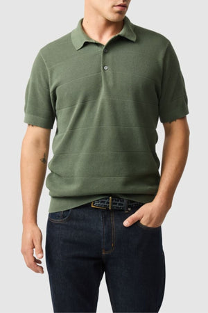 A man wearing a Rodd & Gunn Masons Flat Knit olive green cotton polo and dark blue jeans stands with one hand in his pocket against a plain light gray background.