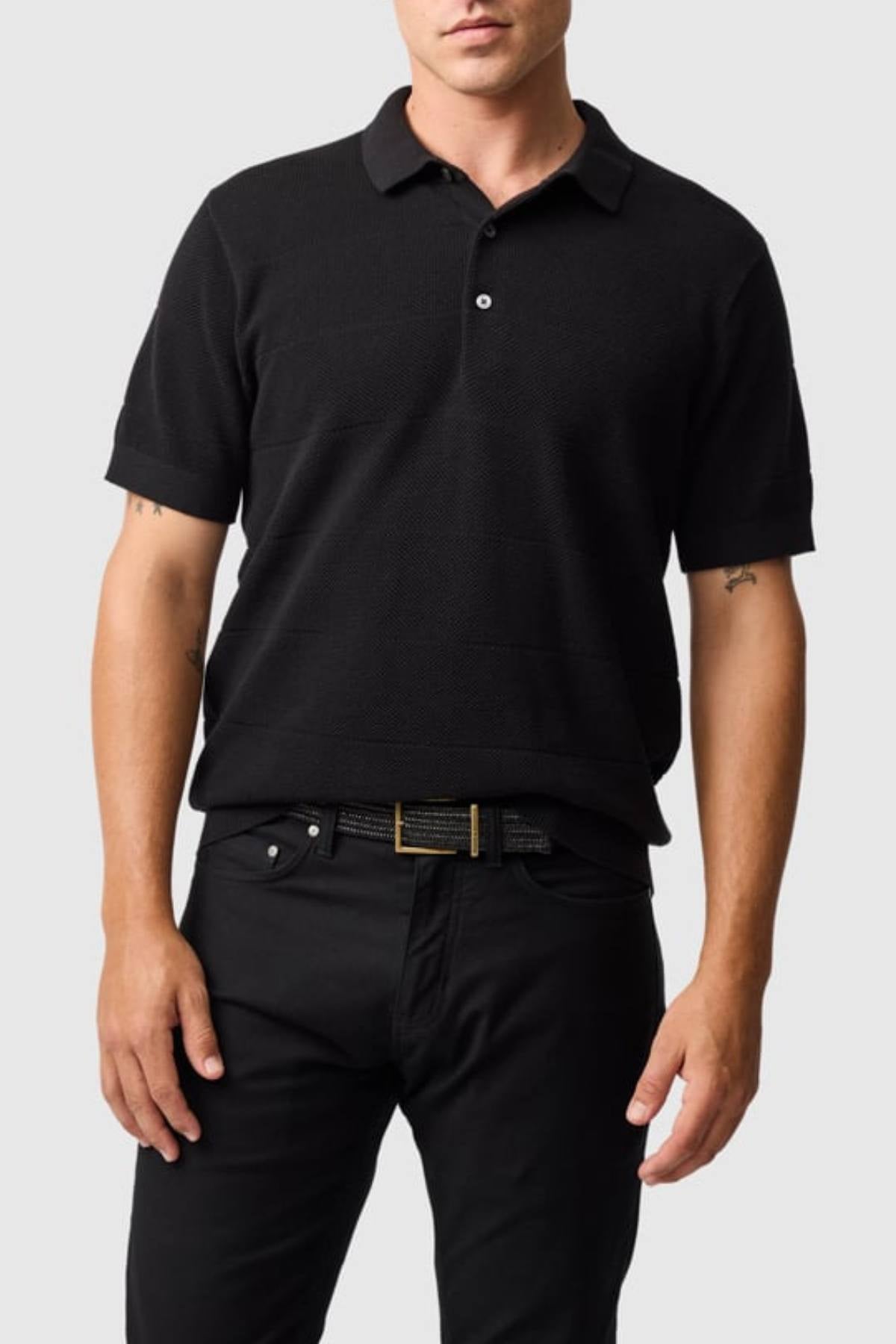 A man wears the Rodd & Gunn Masons Flat Knit black cotton pique shirt and black pants with a belt, standing against a plain light gray background. His arms are relaxed at his sides, and his face is partially visible.