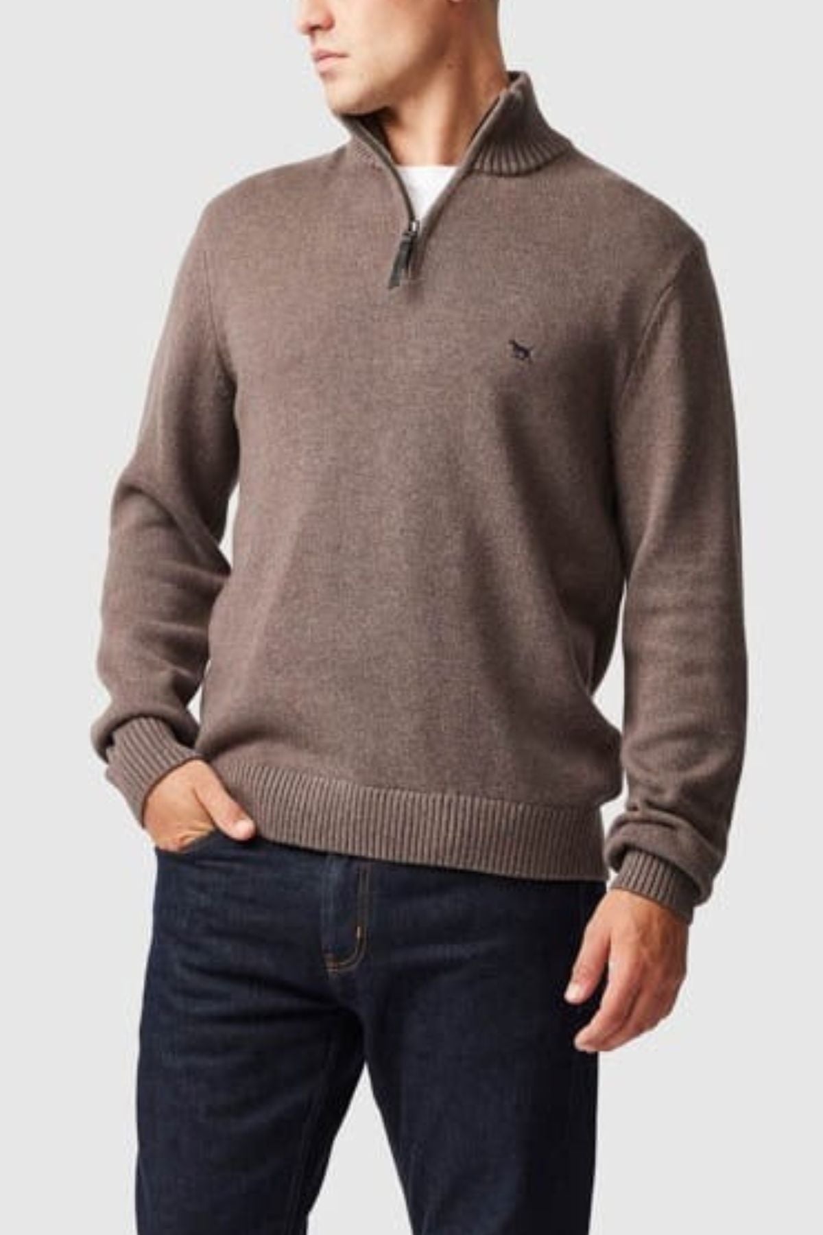 A man wearing the Rodd & Gunn Merrick Bay Knit, a 100% cotton quarter-zip sweater with ribbed trims, pairs it with dark blue jeans and stands with one hand in his pocket against a light gray background.