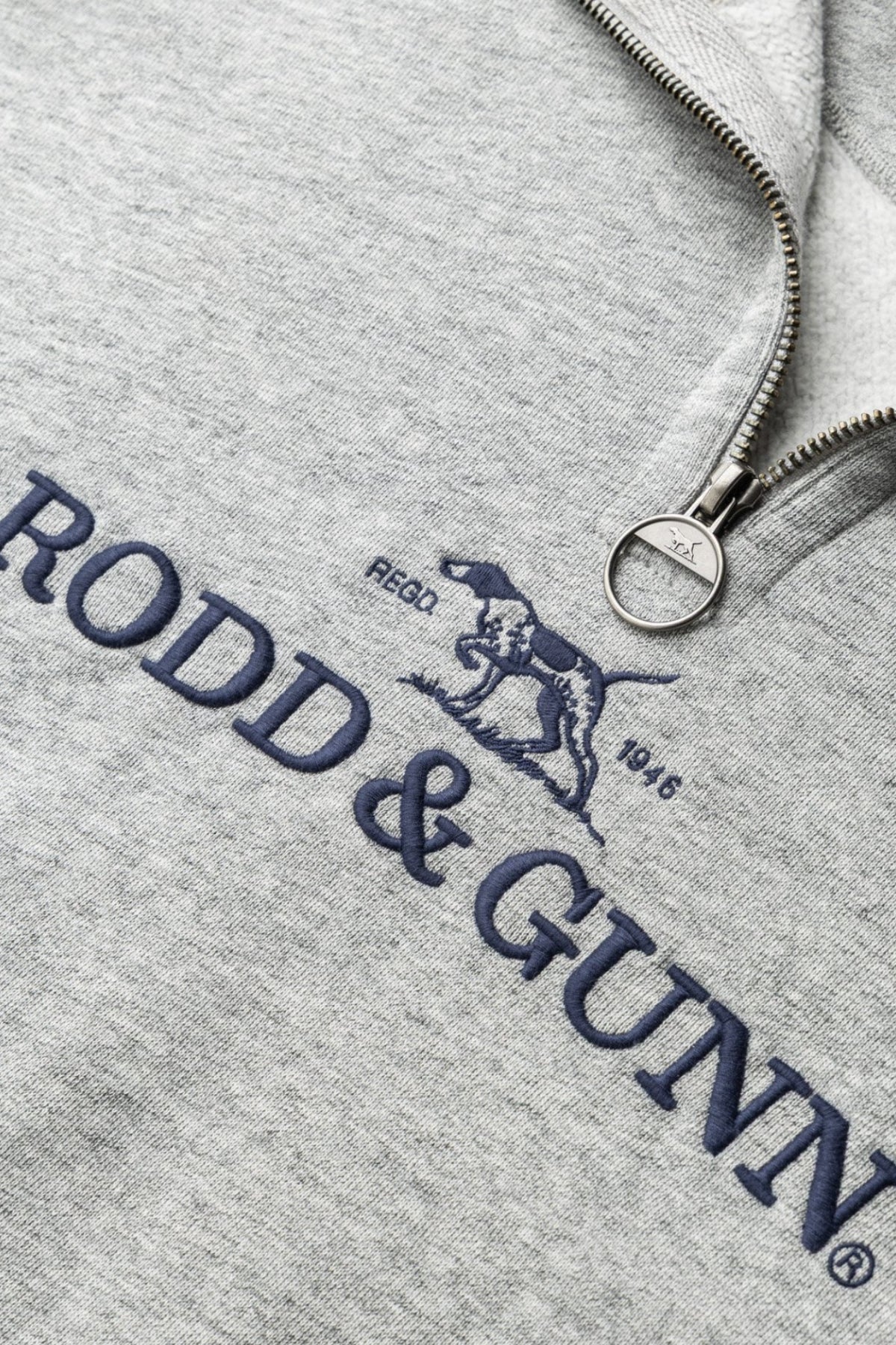 Close-up of the Rodd & Gunn Milford Sound Sweat in light gray, highlighting a premium YKK metal zip, navy "RODD & GUNN" logo, embroidered dog graphic, and "RECO. 1946" text above the logo.