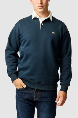 A man in a Rodd & Gunn Otago Sweat over a white collared shirt and dark jeans stands with one hand in his pocket against a plain light gray background.