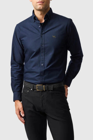 A man stands with one hand in his pocket, wearing a dark blue Rodd & Gunn Gunn Oxford Sports Fit Shirt with long sleeves, black pants, and a black belt against a plain light gray background.