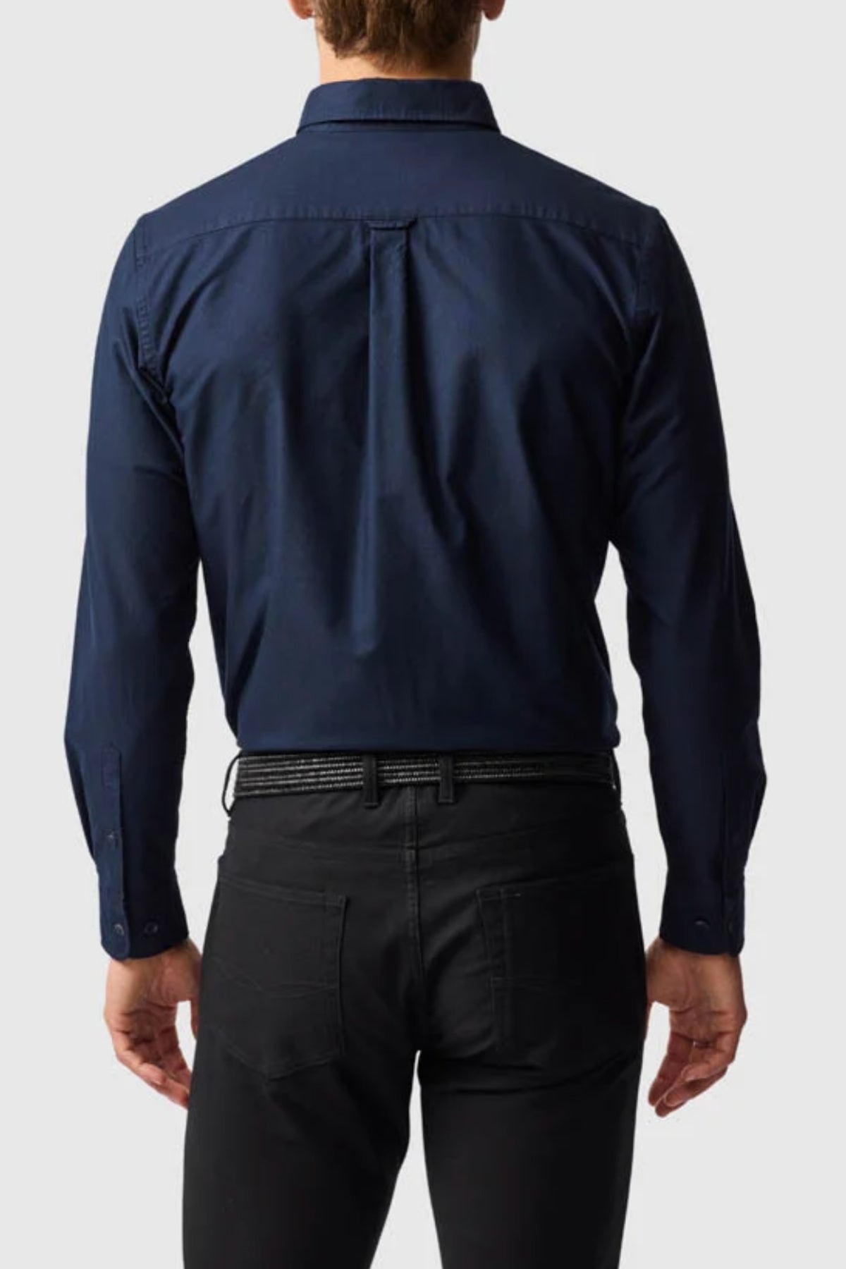 A man is seen from the back wearing a dark blue Rodd & Gunn Gunn Oxford Sports Fit Shirt with black pants and a black belt, standing against a plain light grey background.