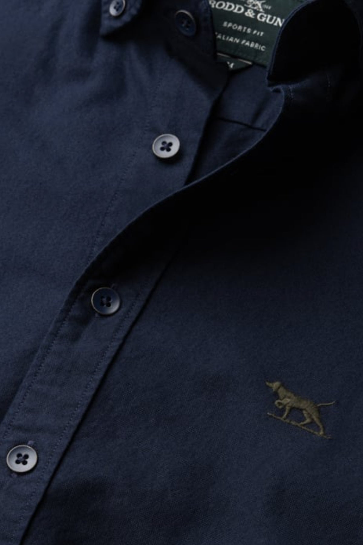 A close-up of the Rodd & Gunn Gunn Oxford Sports Fit Shirt in navy blue, showing a small embroidered dog logo on the chest and part of the inside collar label. Made from 100% cotton for comfort and style.