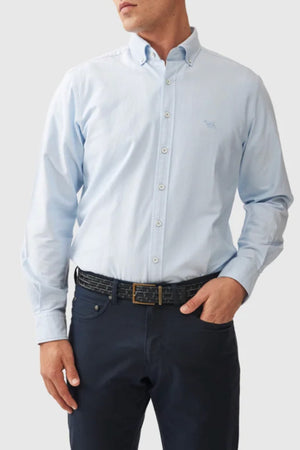 A man wears the Rodd & Gunn Gunn Oxford Sports Fit Shirt in light blue with long sleeves, tucked into dark navy pants. He pairs it with a black belt with a gold buckle, one hand in his pocket, against a plain gray background.
