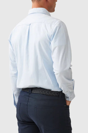 A man stands with his back to the camera, wearing a light blue Rodd & Gunn Gunn Oxford Sports Fit Shirt tucked into dark blue pants with a patterned belt, left hand in pocket, against a plain light gray background.