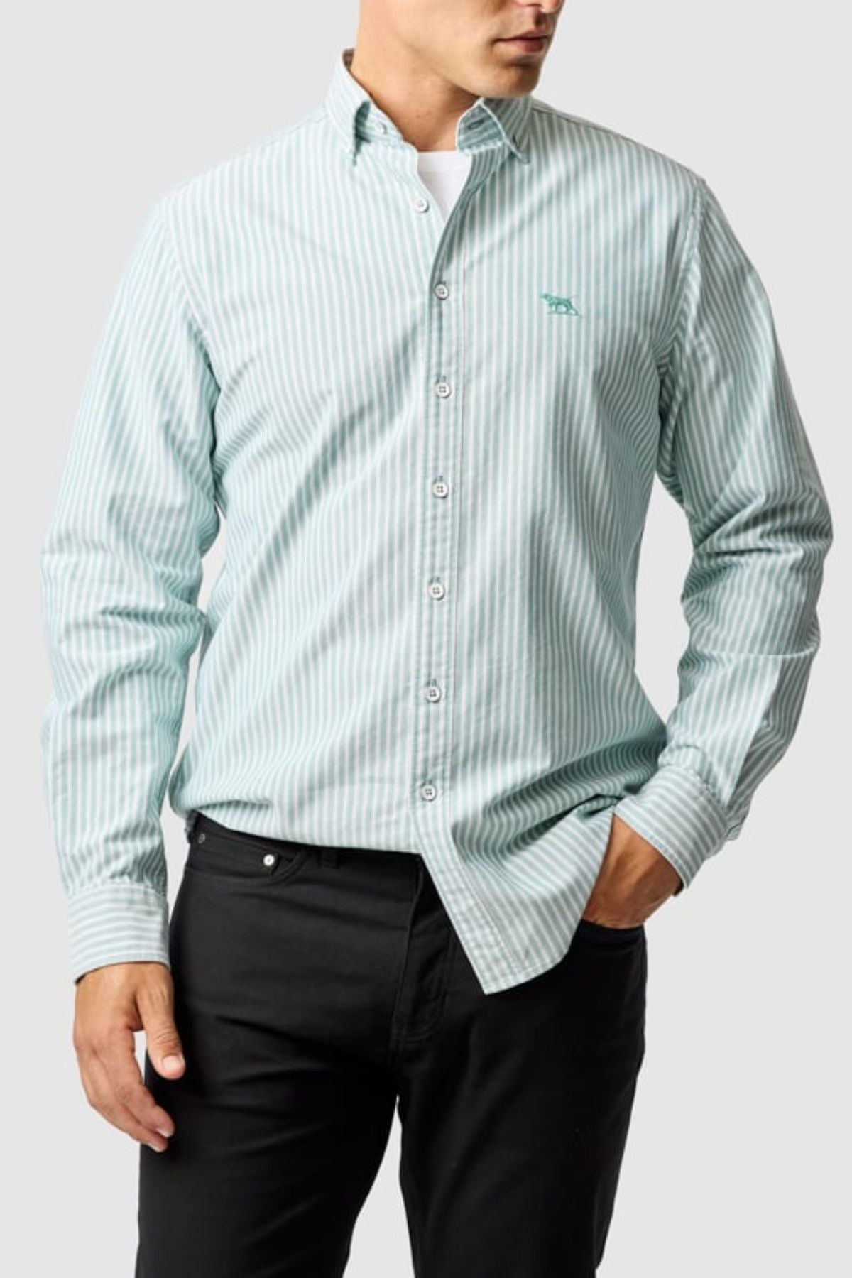 A man wears the Rodd & Gunn Gunn Oxford Stripe 4 Of Shirt in light green and white with long sleeves, paired with black pants, standing with one hand in his pocket against a plain light background.