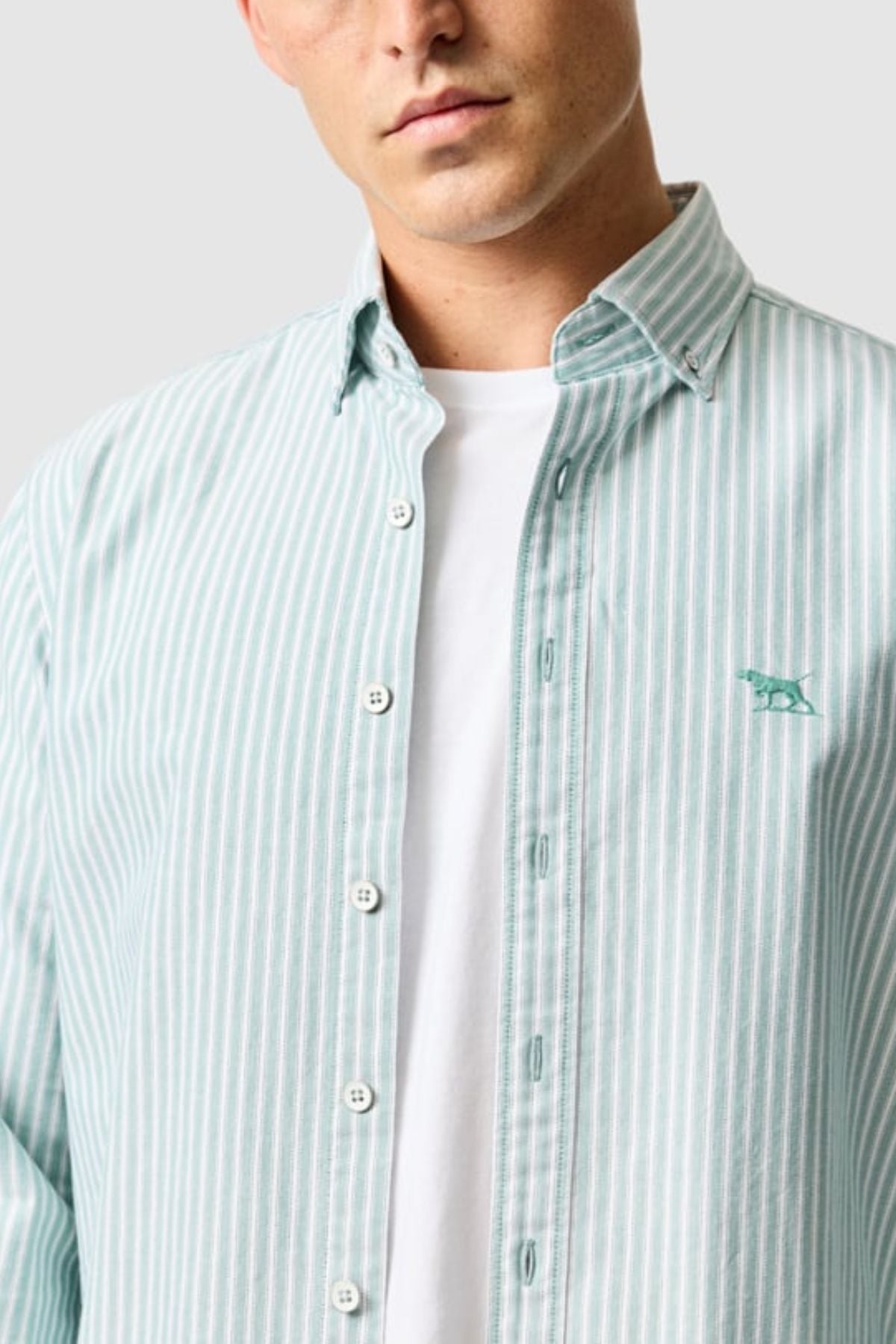 A man wears the Rodd & Gunn Gunn Oxford Stripe 4 Of Shirt in light green and white over a plain white tee. The shirt features a button-down collar and a small animal logo. His face is partially visible.