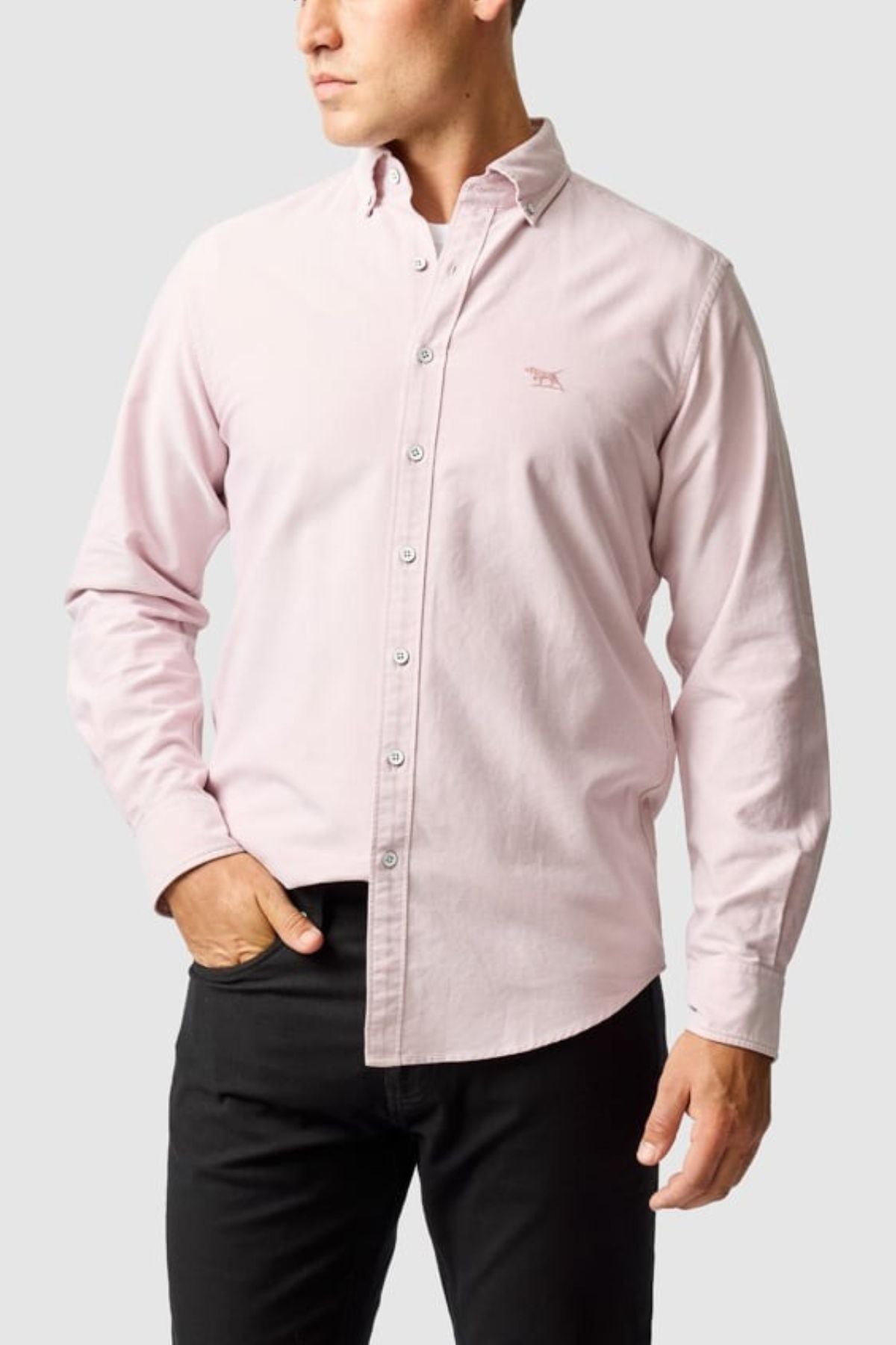 A man wears the Rodd & Gunn Gunn Oxford Sports Fit Shirt in light pink with long sleeves and black pants, standing with one hand in his pocket against a plain light gray background.