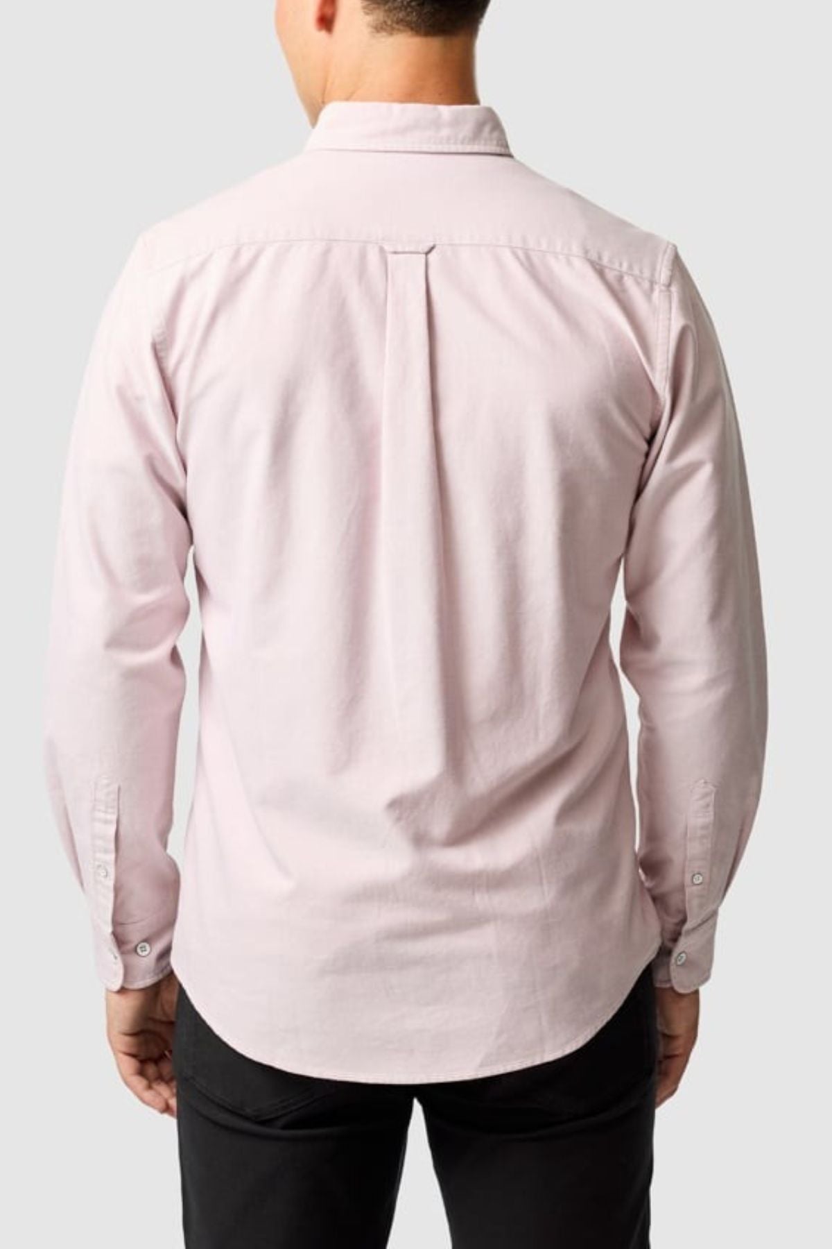 A person is shown from the back wearing the Rodd & Gunn Gunn Oxford Sports Fit Shirt in light pink, made from 100% cotton. This long-sleeve button-up has a center box pleat and buttoned cuffs, set against a plain light gray background.