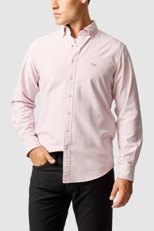 A man wears the Rodd & Gunn Gunn Oxford Sports Fit Shirt in light pink with long sleeves and black pants, standing with one hand in his pocket against a plain light gray background.