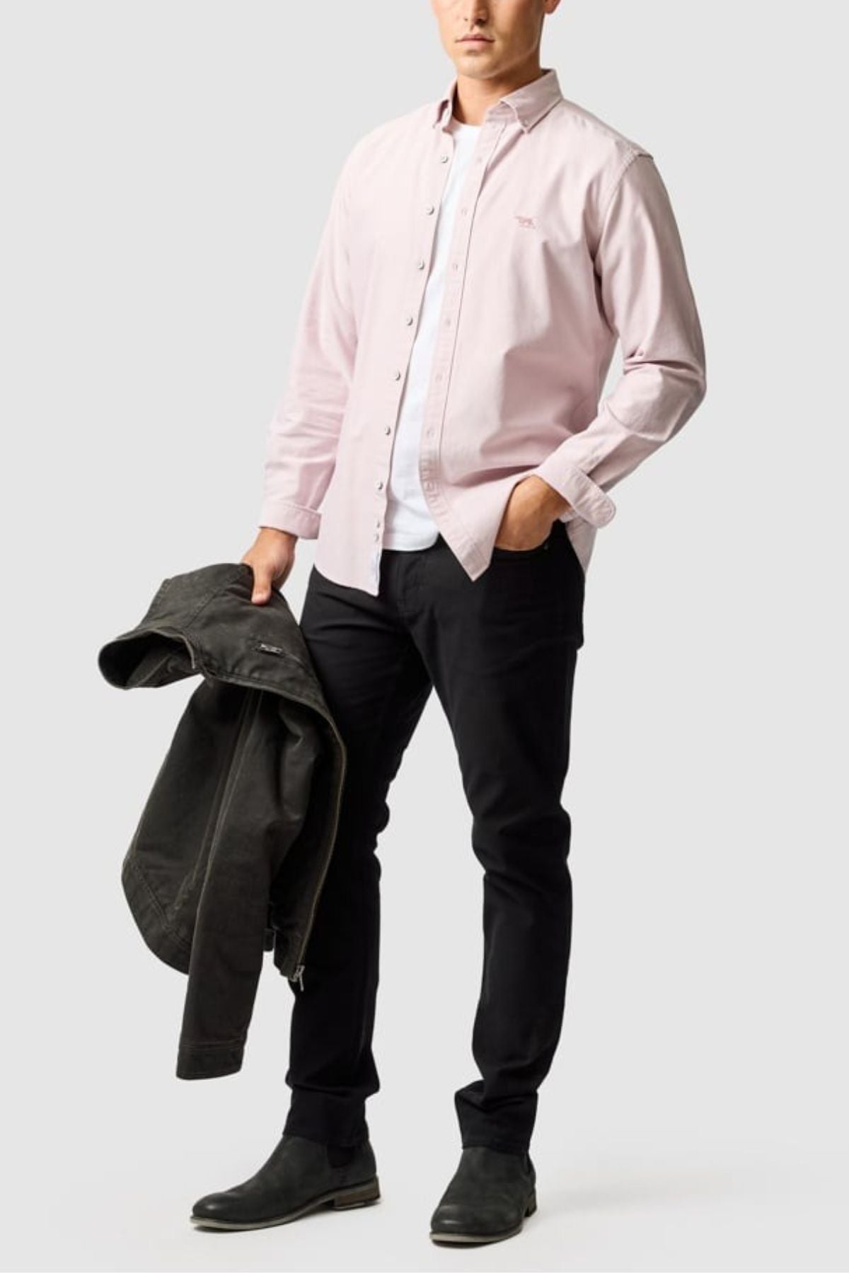 A man stands against a light background, wearing a light pink Rodd & Gunn Gunn Oxford Sports Fit Shirt (100% cotton), white t-shirt, black jeans and shoes, and holding a dark jacket in his left hand.
