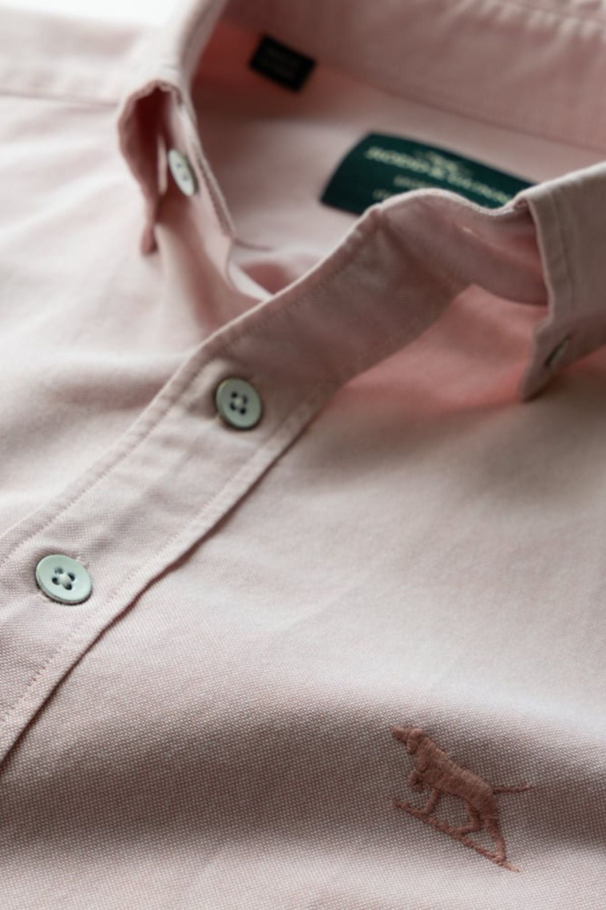 A close-up of the Rodd & Gunn Gunn Oxford Sports Fit Shirt in pale pink, made from 100% cotton, with white buttons and a small embroidered animal logo on the front. The shirt label is partially visible inside the collar.