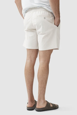 A man stands with his hand in his pocket, wearing Rodd & Gunn Resort Short in a light color, paired with a white shirt and dark sandals, shown from the back against a plain background.