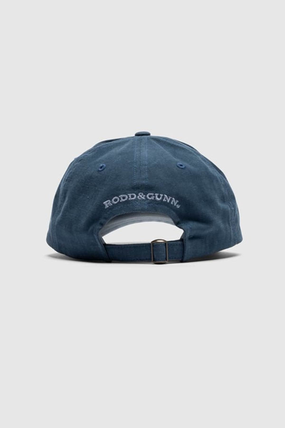 The Rodd & Gunn Signature Cap in blue is displayed from the back, highlighting its adjustable strap with a metal buckle and white "RODD & GUNN" embroidery. Made from cotton twill, it is set against a light gray background.