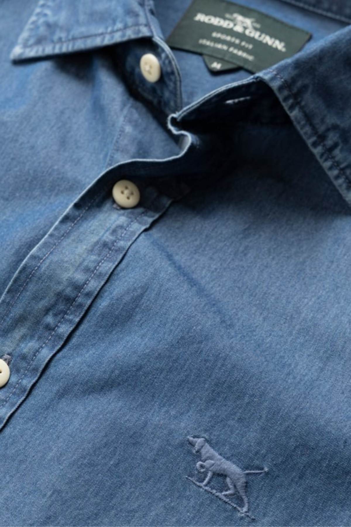 Close-up of a blue Rodd & Gunn Spring Creek Sports Fit Shirt with white buttons and an embroidered dog logo near the hem. The visible label reads "Rodd & Gunn, Linen Shirt." This sports fit shirt blends timeless style with attention to detail.