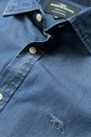 Close-up of a blue Rodd & Gunn Spring Creek Sports Fit Shirt with white buttons and an embroidered dog logo near the hem. The visible label reads "Rodd & Gunn, Linen Shirt." This sports fit shirt blends timeless style with attention to detail.