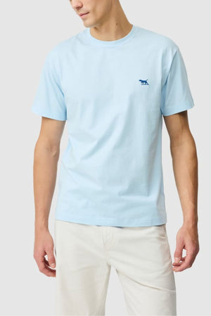 A man stands against a plain light gray background wearing the Rodd & Gunn The Gunn Tee 2.0—a light blue 100% cotton sports fit T-shirt with a small blue dog logo on the left chest—paired with light beige pants.