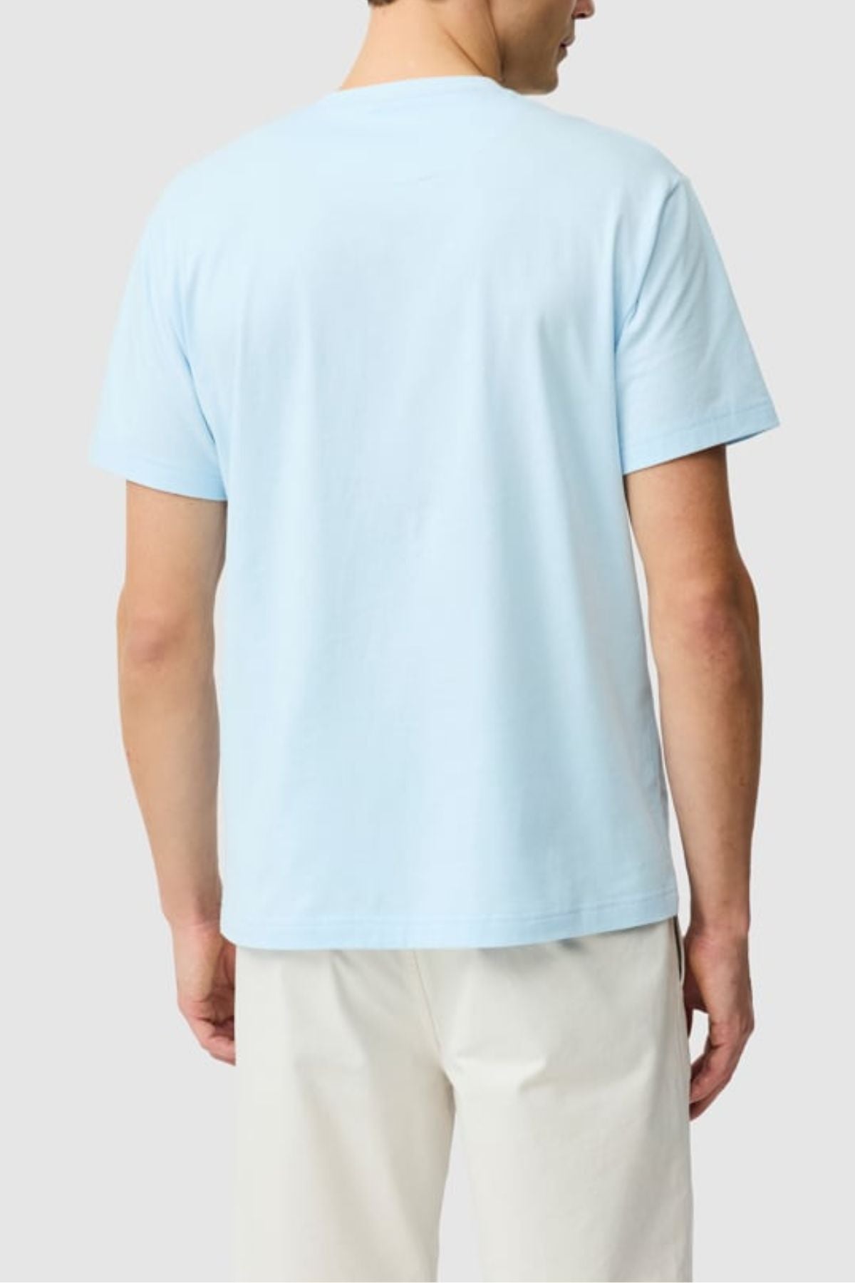 A person faces away from the camera, wearing the Rodd & Gunn The Gunn Tee 2.0 in light blue (100% cotton) and off-white pants, set against a plain light gray background.
