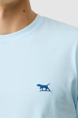 A person wears the Rodd & Gunn The Gunn Tee 2.0 in light blue, with a small, dark blue embroidered dog logo on the chest. Only the round neckline and part of the neck and shoulder are visible.