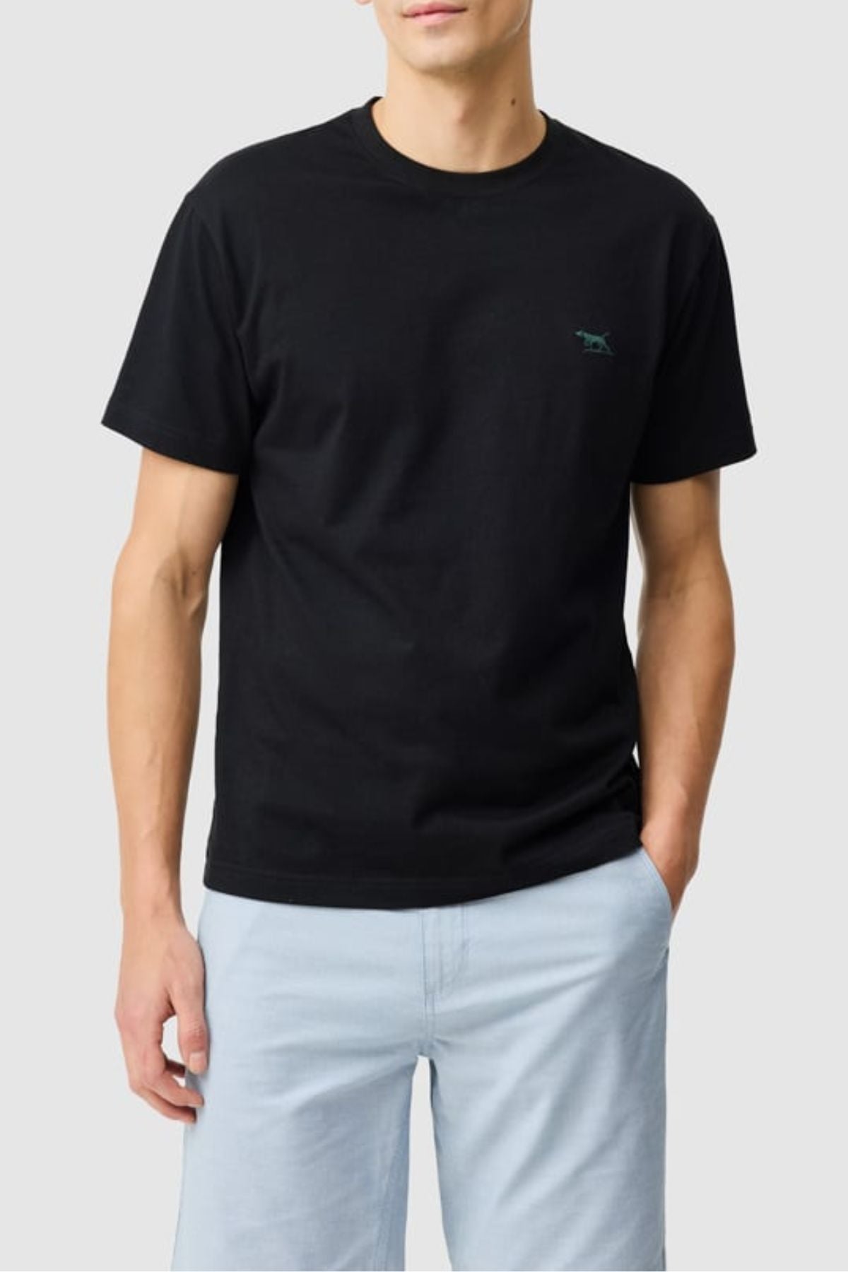 A person wears the Rodd & Gunn The Gunn Tee 2.0—a plain black 100% cotton tee with a small chest logo—paired with light blue shorts, standing with one hand in their pocket against a light gray background.