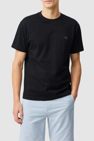 A person wears the Rodd & Gunn The Gunn Tee 2.0—a plain black 100% cotton tee with a small chest logo—paired with light blue shorts, standing with one hand in their pocket against a light gray background.