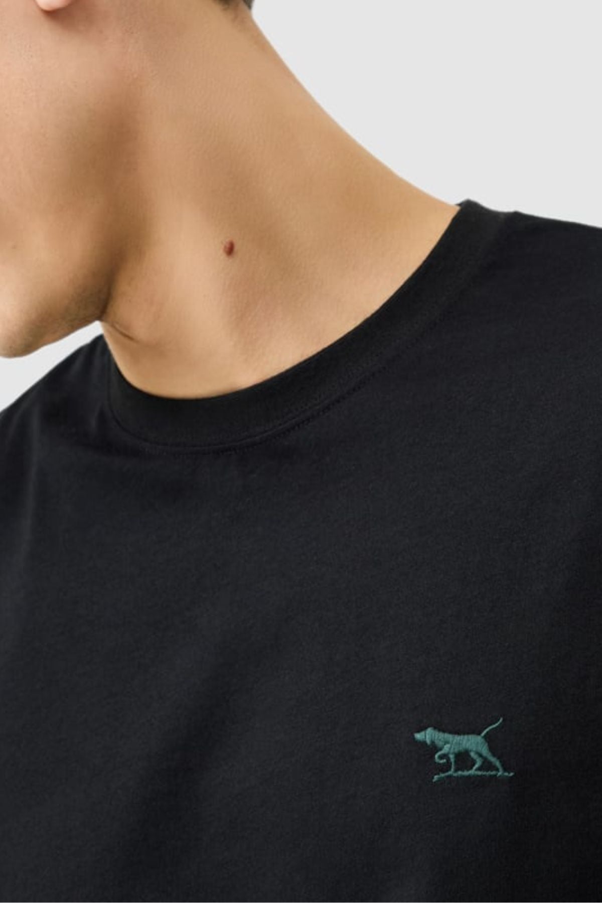 A person wearing the Rodd & Gunn The Gunn Tee 2.0 in black, featuring a small green embroidered dog logo on the chest; only their lower face, neck, and upper torso are visible.