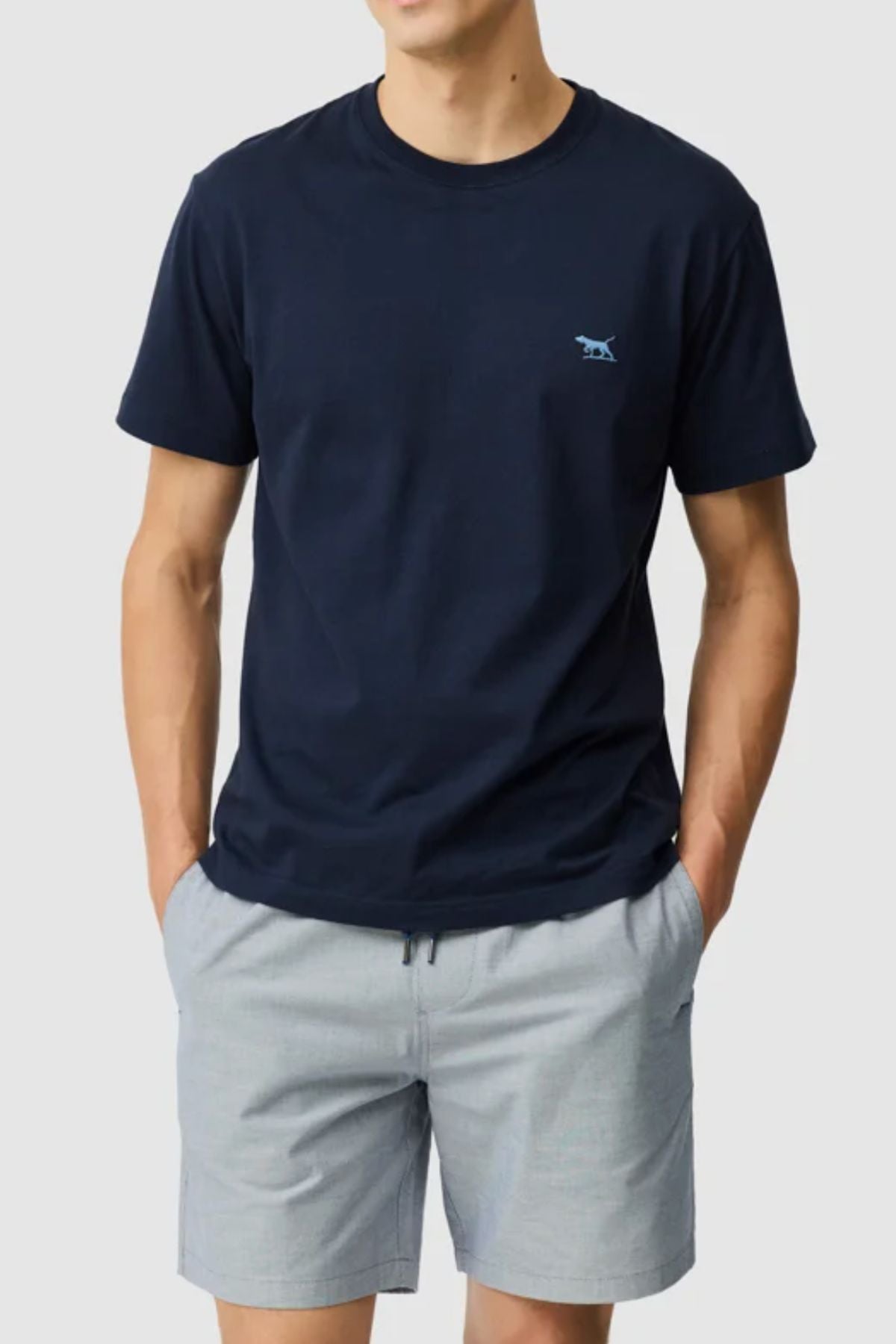 A person stands with hands in pockets against a light background, wearing the Rodd & Gunn The Gunn Tee 2.0— a dark navy men’s 100% cotton crew neck t-shirt with a small embroidered animal logo—paired with light gray shorts.