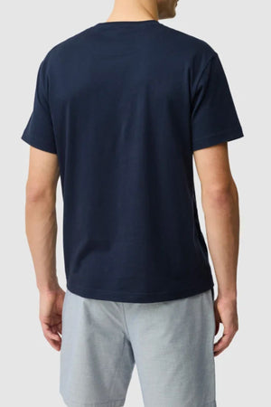 A person is shown from the back, wearing a Rodd & Gunn The Gunn Tee 2.0 in plain navy blue and light gray shorts, standing against a light gray background.
