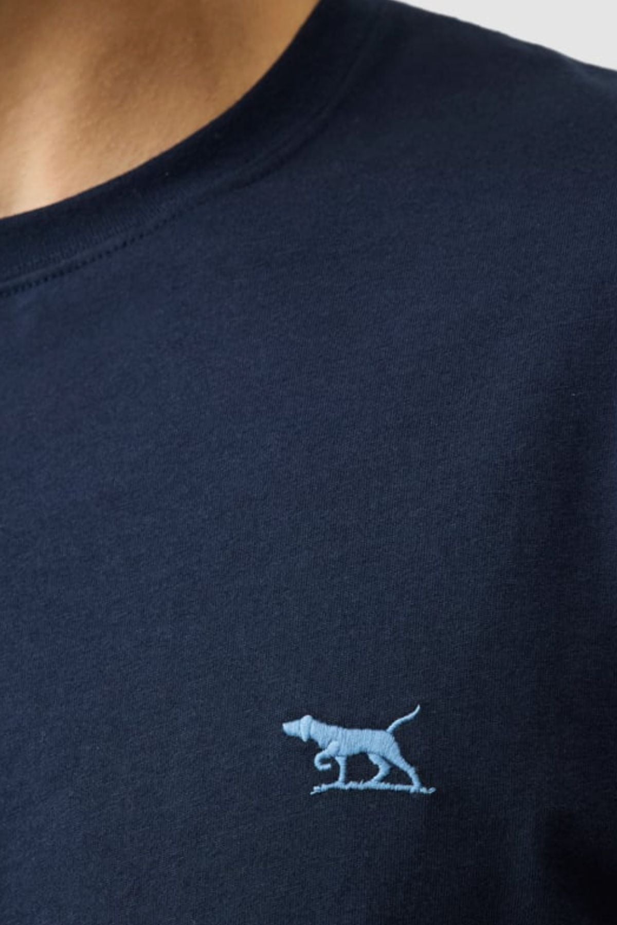 Close-up of a person wearing the Rodd & Gunn The Gunn Tee 2.0 in navy blue, featuring a small light blue embroidered dog logo on the left chest and made from 100% cotton with a classic crew neck.