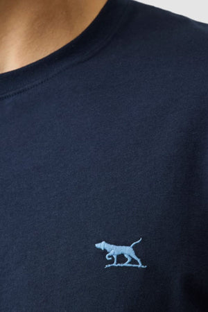 Close-up of a person wearing the Rodd & Gunn The Gunn Tee 2.0 in navy blue, featuring a small light blue embroidered dog logo on the left chest and made from 100% cotton with a classic crew neck.