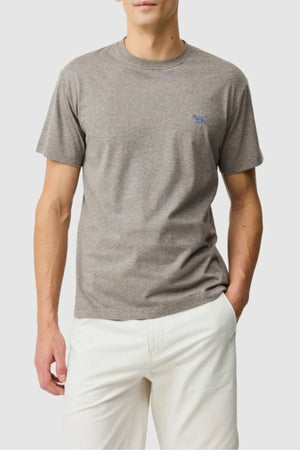 Someone wears the Rodd & Gunn The Gunn Tee 2.0 in light brown, crafted from breathable jersey fabric, paired with white shorts, standing with one hand in their pocket against a plain light background.
