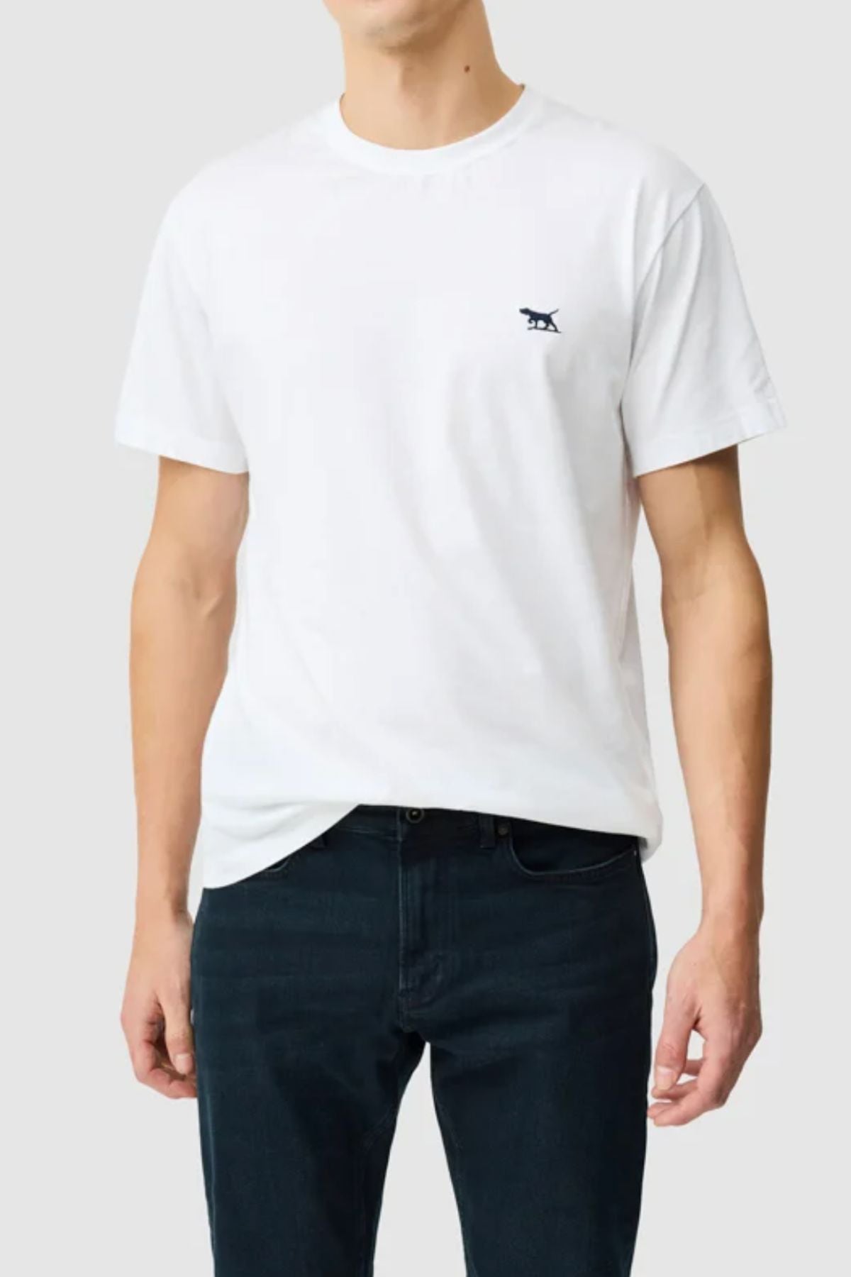 A person wears the Rodd & Gunn The Gunn Tee 2.0—a plain white sports fit jersey t-shirt with a small black dog logo on the left chest—paired with dark jeans, standing against a light gray background.
