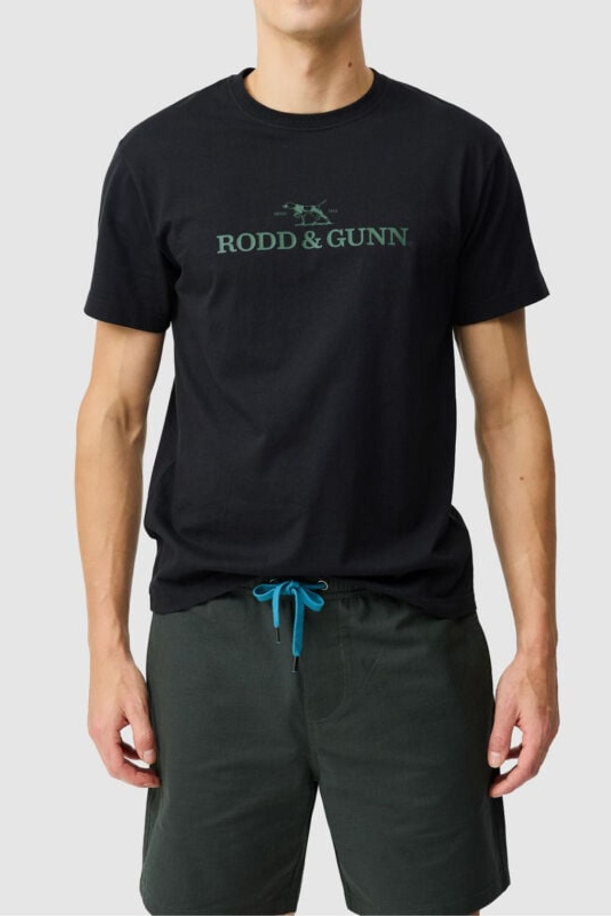 A man stands against a light grey background wearing the Rodd & Gunn The Logo Tee 2.0, made from 100% cotton, paired with dark green shorts featuring blue drawstrings.