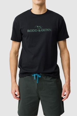A man stands against a light grey background wearing the Rodd & Gunn The Logo Tee 2.0, made from 100% cotton, paired with dark green shorts featuring blue drawstrings.