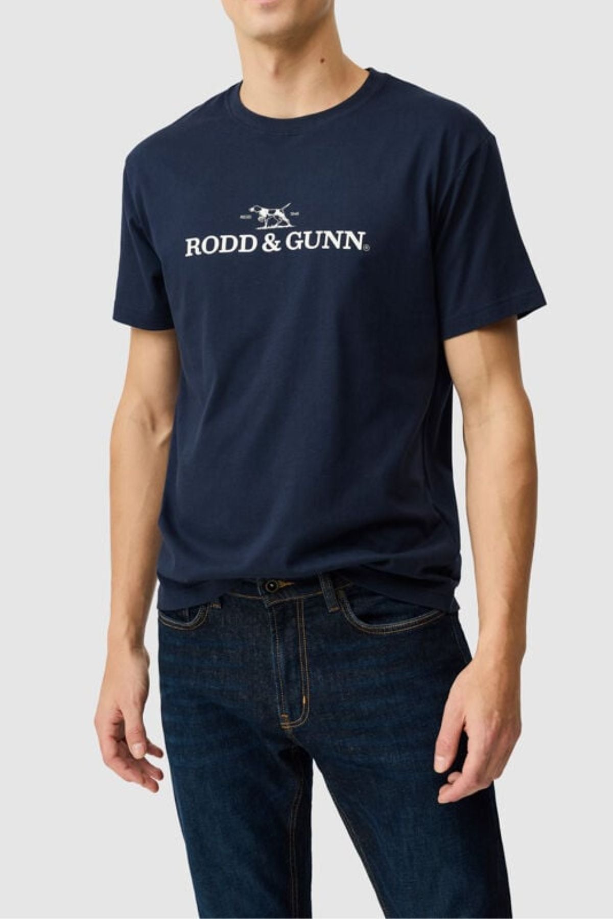 A man wears the Rodd & Gunn The Logo Tee 2.0 in navy blue, featuring white text and a small logo on 100% cotton, paired with dark blue jeans, standing against a plain light gray background.