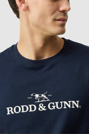 A man wears the Rodd & Gunn The Logo Tee 2.0, a navy sports fit t-shirt featuring a white dog graphic and "RODD & GUNN" on the front, crafted from 100% cotton for comfort.
