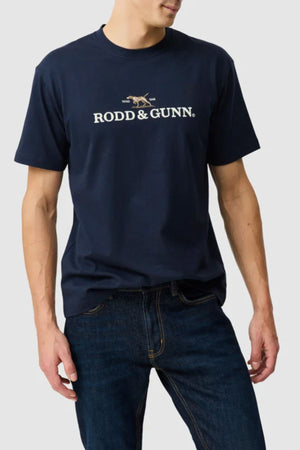 A man wears the Rodd & Gunn The Peaks Logo Tee, featuring FibreTrace technology and a crossed rifles graphic, with dark blue jeans against a light gray background.