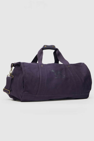 The Rodd & Gunn Ponsonby Road Duffle Bag, crafted from dark purple cotton canvas, features short handles, a detachable shoulder strap, and a subtle side logo, displayed against a plain light background.