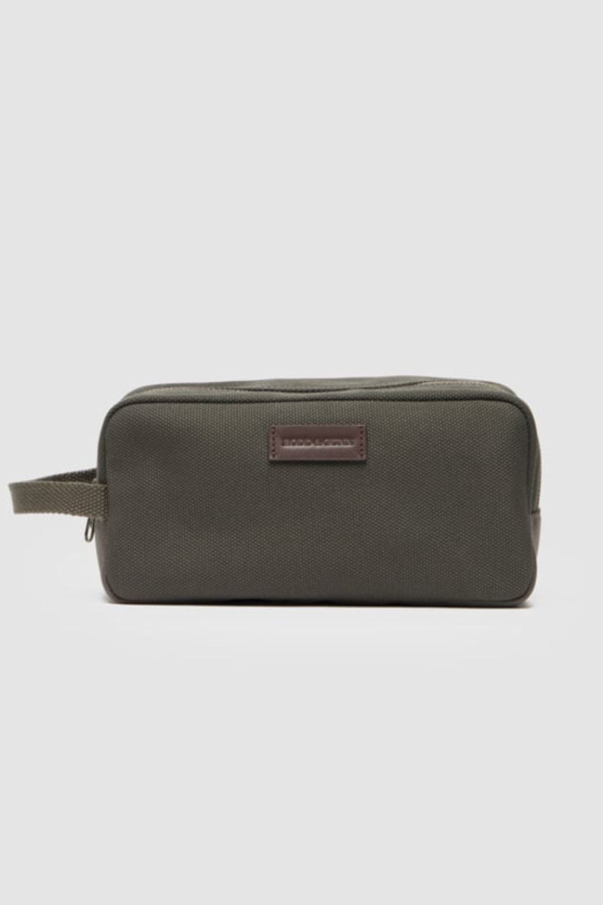 The Rodd & Gunn Ponsonby Wash Bag is a rectangular olive green cotton canvas bag with a side handle and a small leather patch on the front, shown against a plain light gray background.