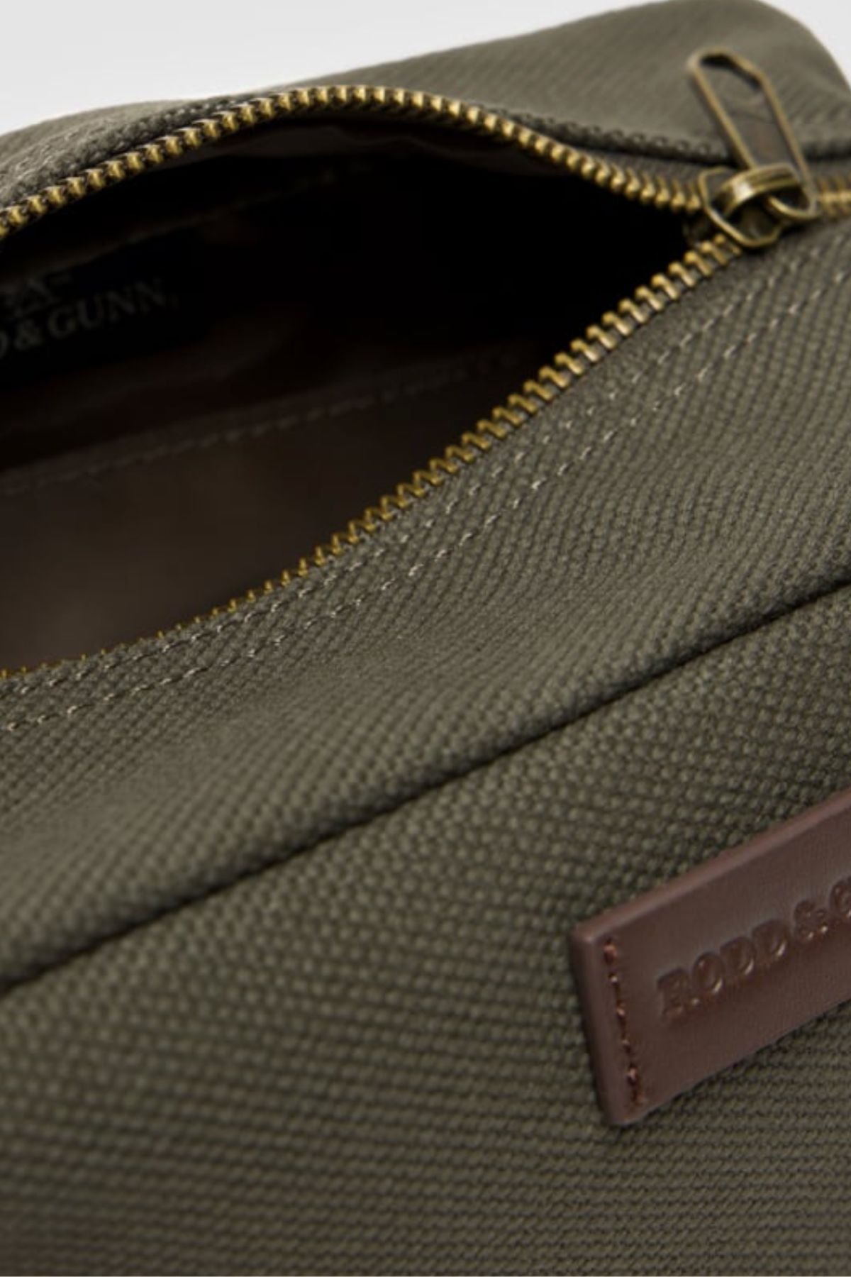 A close-up of the Rodd & Gunn Ponsonby Wash Bag, crafted from olive green cotton canvas with durable textured fabric, a partially open brass zipper, and a brown leather patch label embossed with text.