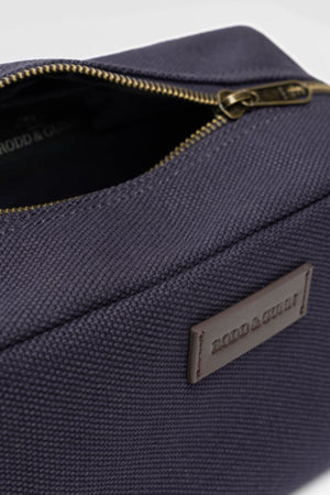 A close-up of the Rodd & Gunn Ponsonby Wash Bag shows its dark navy textured cotton canvas, brass zipper, and front leather patch labeled "RODD & GUNN.