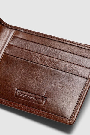 A close-up of an open Rodd & Gunn Wakefield Bi-Fold Wallet made from brown Italian leather with multiple card slots and "MADE IN PORTUGAL" embossed inside. The RFID-protected wallet sits empty on a plain light gray background.