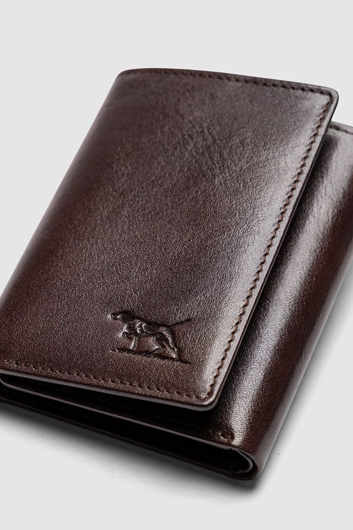 The Rodd & Gunn Westport Tri Fold Wallet is crafted from dark brown Italian leather with a smooth finish and features an embossed dog logo on the lower front, shown against a light gray background.