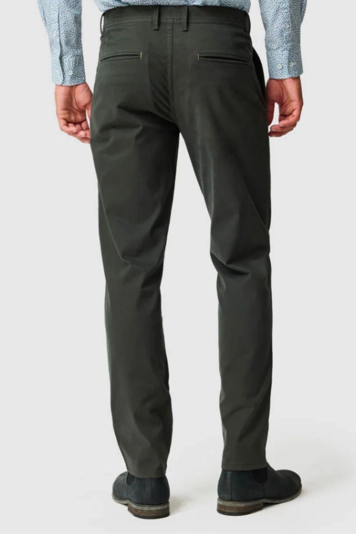 A man stands with his back to the camera against a plain background, wearing dark green Rodd & Gunn Thomas Road 2.0 Chino Pants, a light blue patterned shirt, and black shoes.