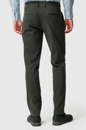 A man stands with his back to the camera against a plain background, wearing dark green Rodd & Gunn Thomas Road 2.0 Chino Pants, a light blue patterned shirt, and black shoes.