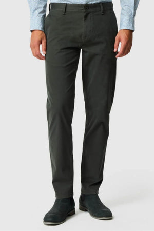 A person wearing Rodd & Gunn Thomas Road 2.0 Chino Pant in dark gray, a light blue patterned dress shirt, and black boots stands against a plain light background. The person's face is not visible.