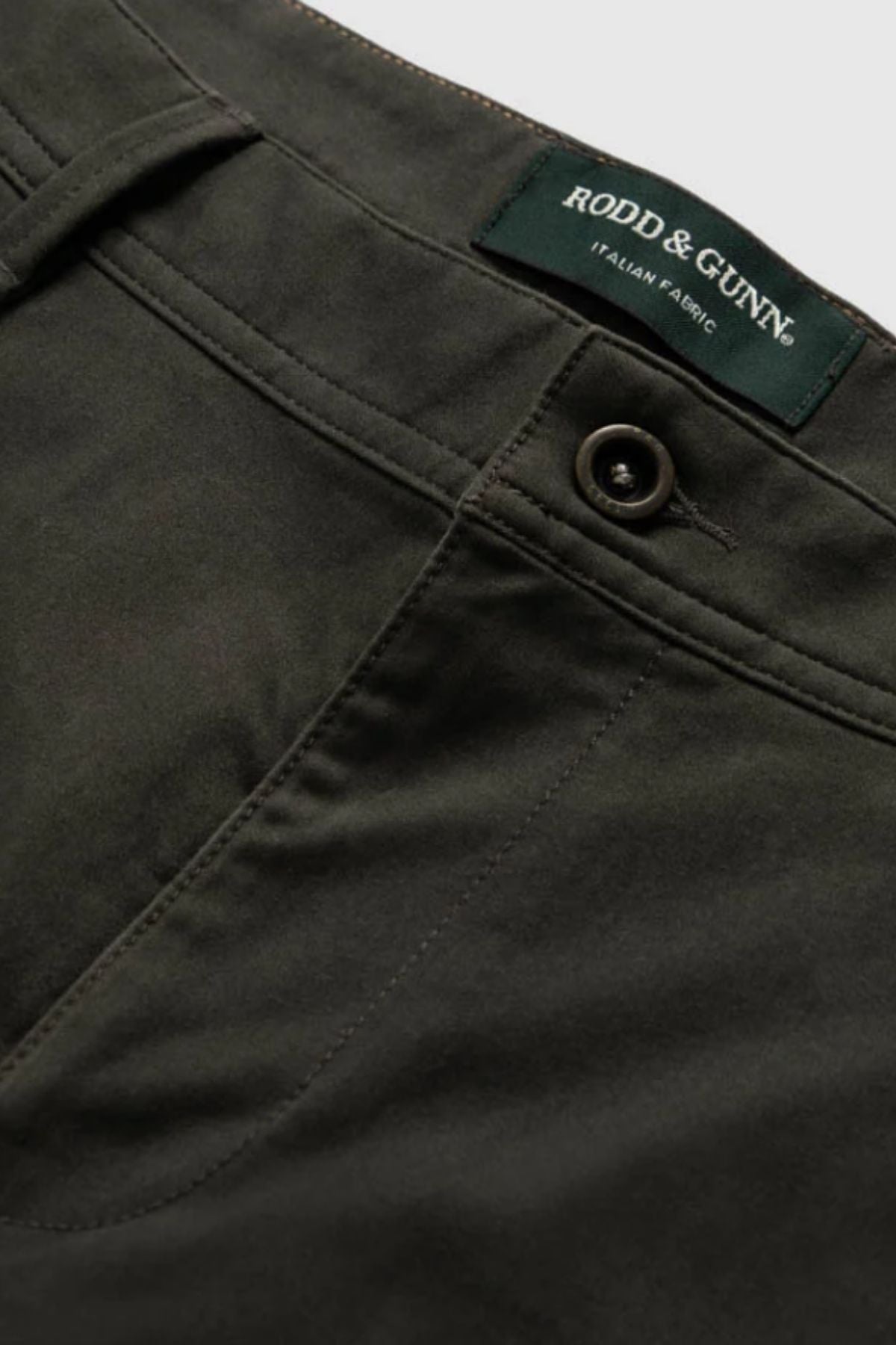 Close-up of Rodd & Gunn Thomas Road 2.0 Chino Pant in dark olive green, featuring a button and pocket detail. The label above the waistband reads "Rodd & Gunn Italian Fabric," highlighting its premium Italian-woven material.