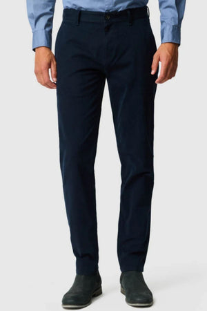 A person wearing a light blue button-up shirt, Rodd & Gunn Thomas Road 2.0 Chino Pant in dark navy, and dark dress shoes stands against a plain light background. Their face is not visible.
